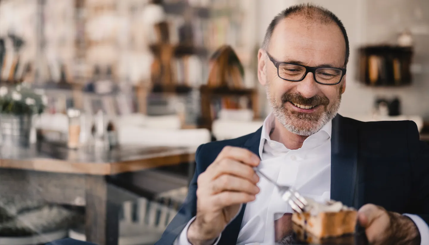 Smiling man in glasses and a suit enjoying a slice of cake at a cozy café.