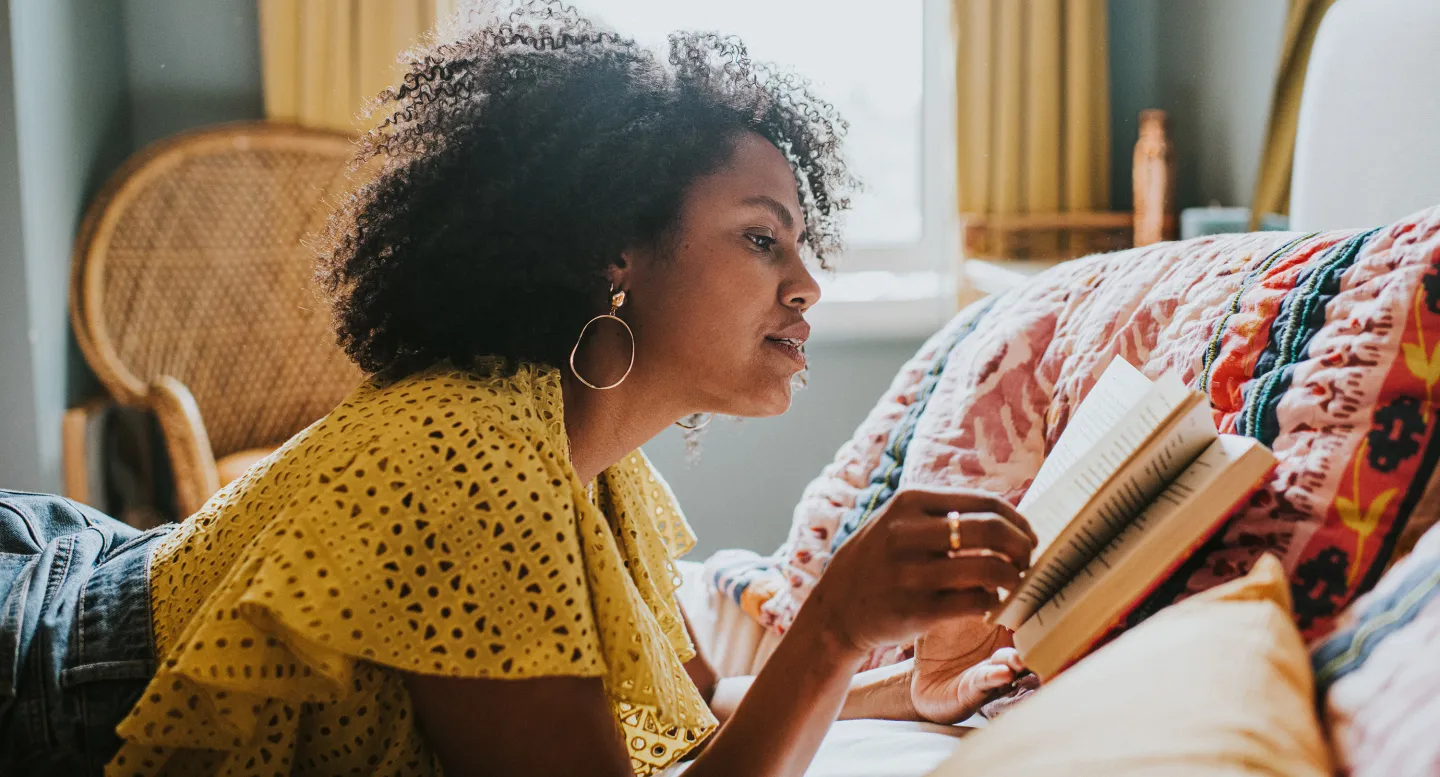 Woman in a yellow top lying on a bed and reading a book in a cozy, sunlit room with colorful bedding.