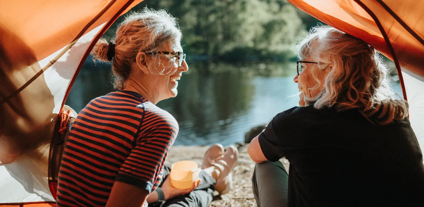  Two older women smiling and talking while sitting at the edge of a tent by a lake, enjoying a peaceful moment in nature.
