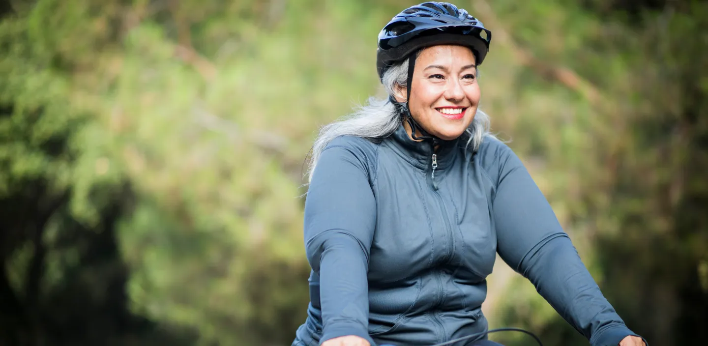 Smiling woman with gray hair wearing a bike helmet and a zip-up jacket, riding a bicycle outdoors.