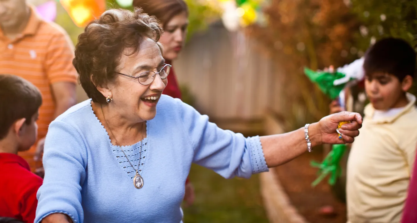Smiling elderly woman in a blue sweater reaching out to children at an outdoor family gathering with colorful decorations.