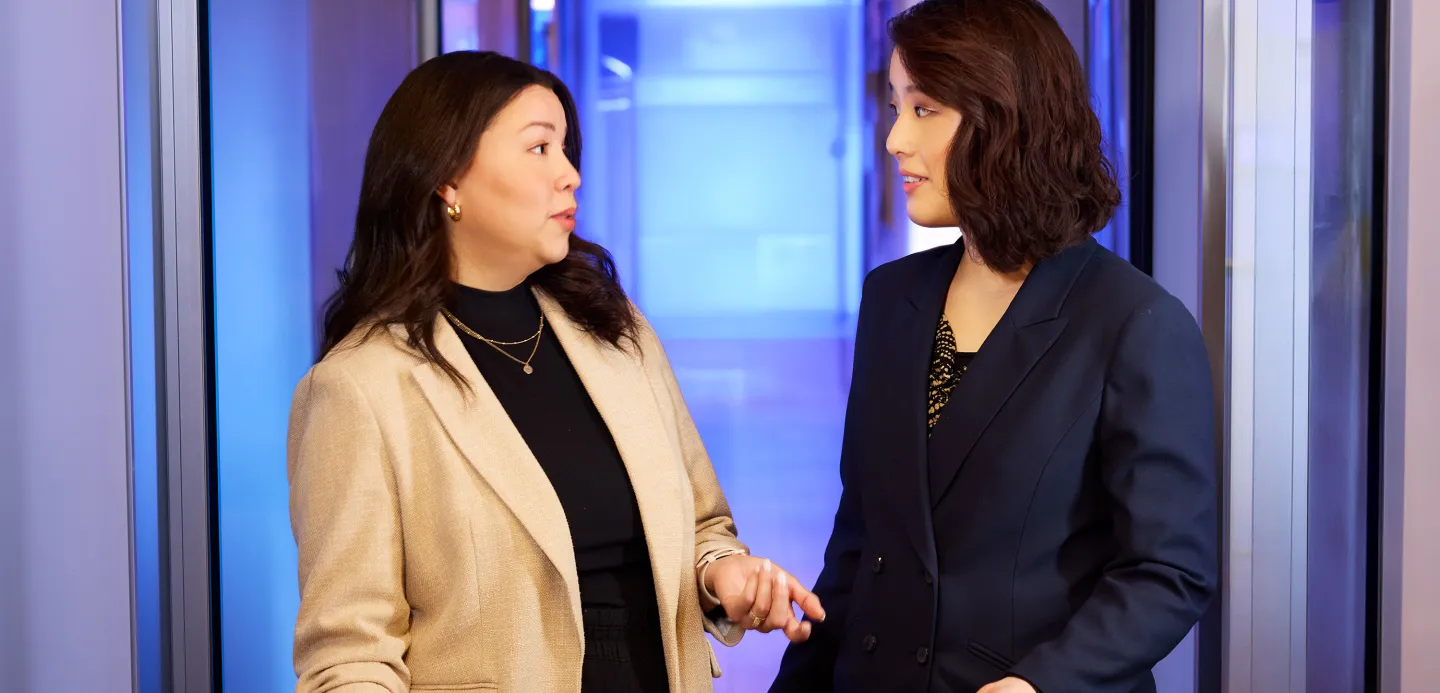 Two women in business attire have a conversation in a brightly lit hallway with blue and purple tones.