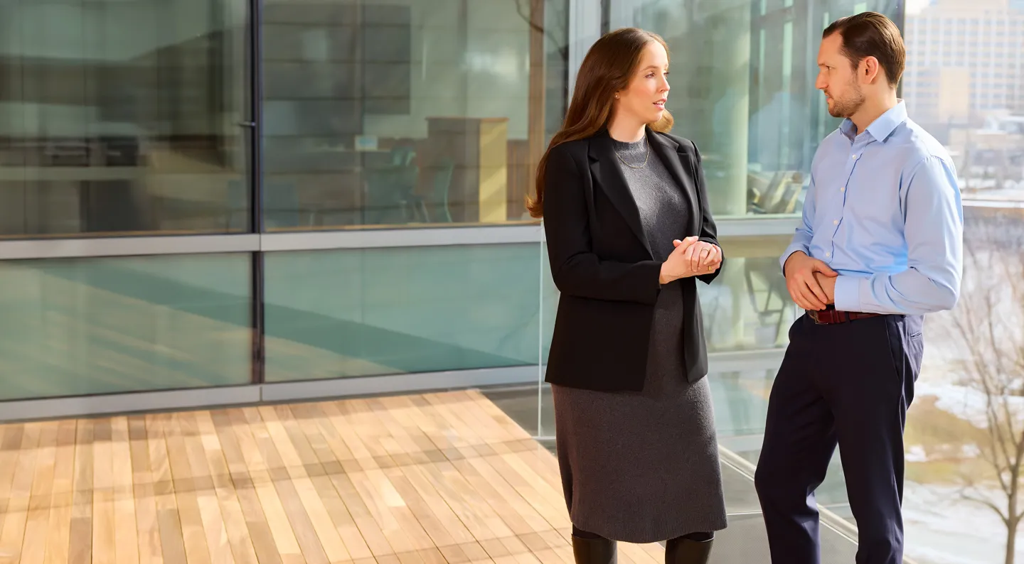 Two professionals talking indoors near a window, woman in black blazer and man in light blue shirt.