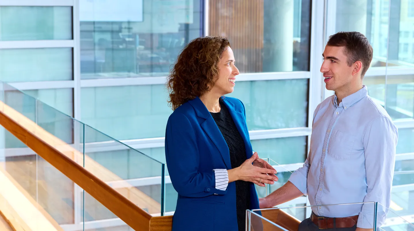 Woman in blue blazer and man in light shirt talking and smiling in a modern glass building.