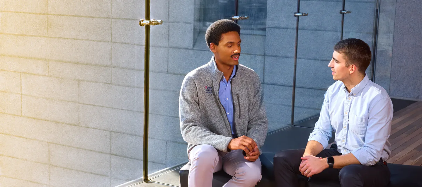 Two men sitting and talking outside against a stone wall, one wearing a Montefiore Einstein fleece.