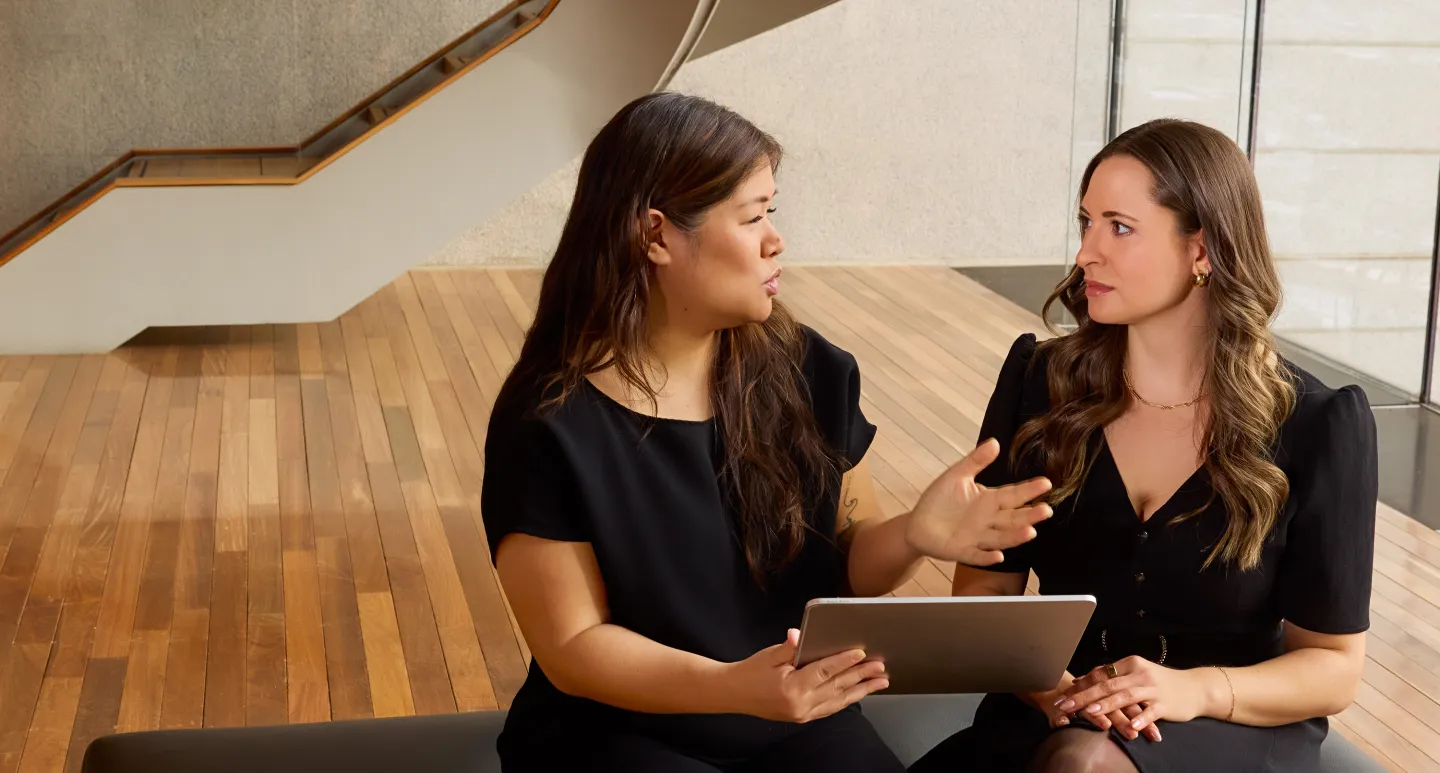 Two women in black outfits sitting and talking on a wooden floor near a staircase and large windows.