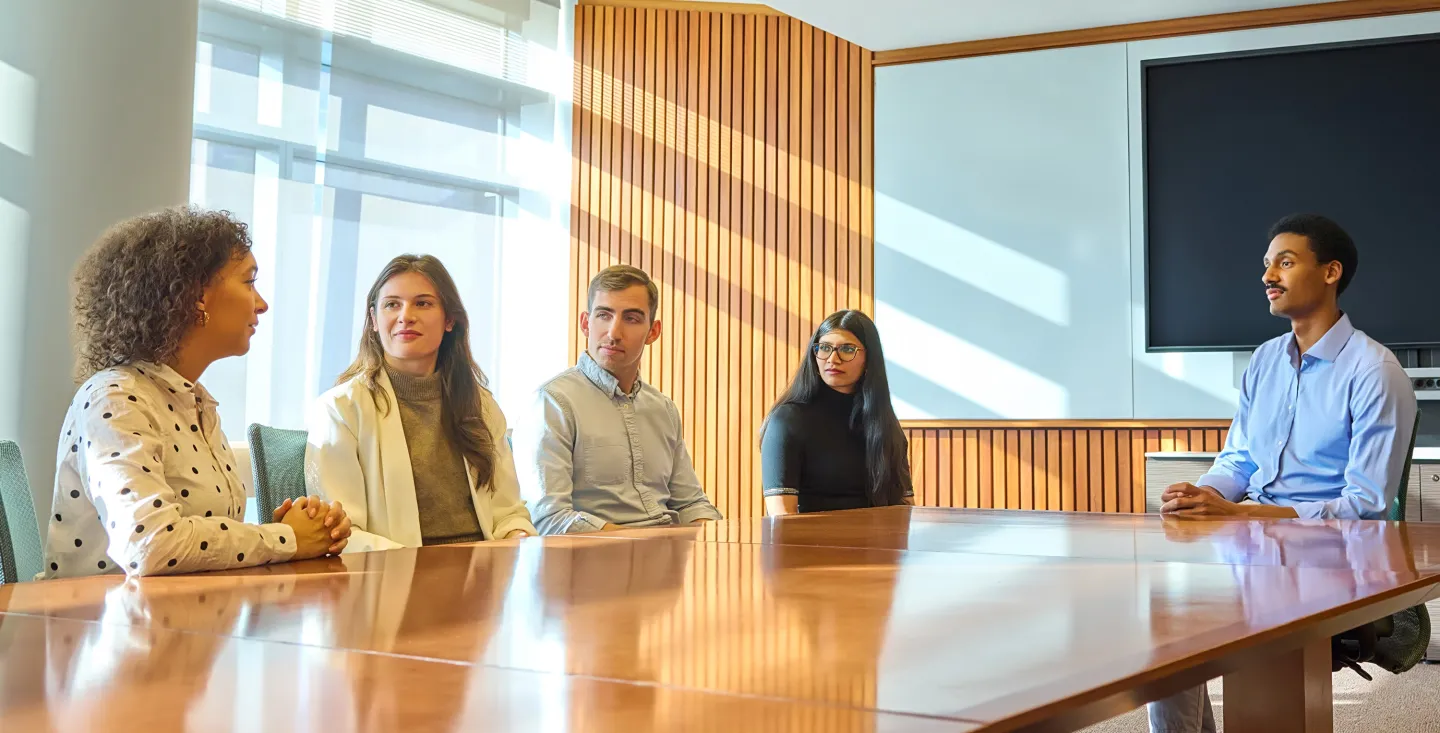 Five colleagues sit around a conference table in a sunlit room, engaged in a discussion with a large screen behind them.