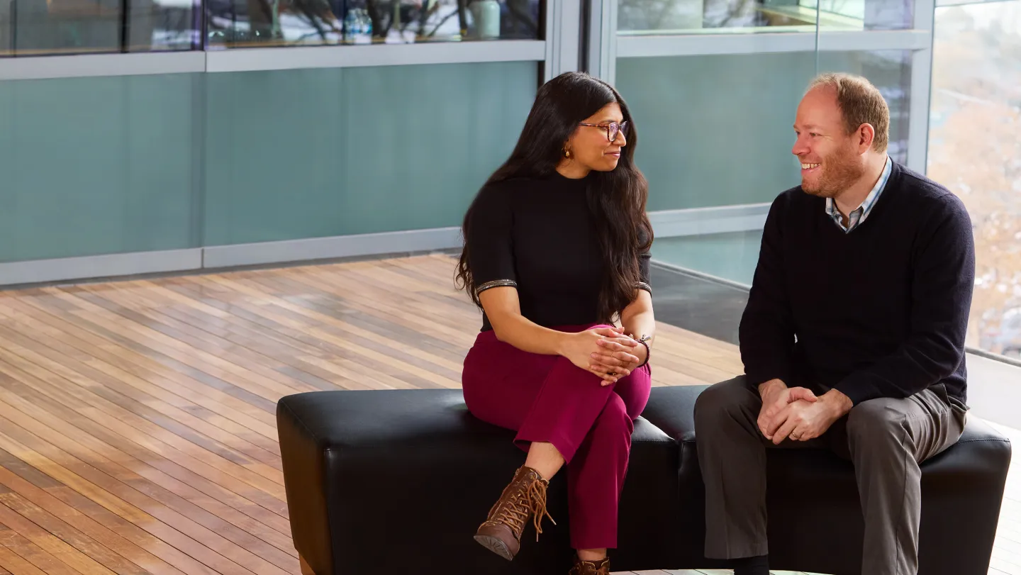 Two colleagues seated and smiling in a conversation near glass walls and a wooden floor inside a modern office.