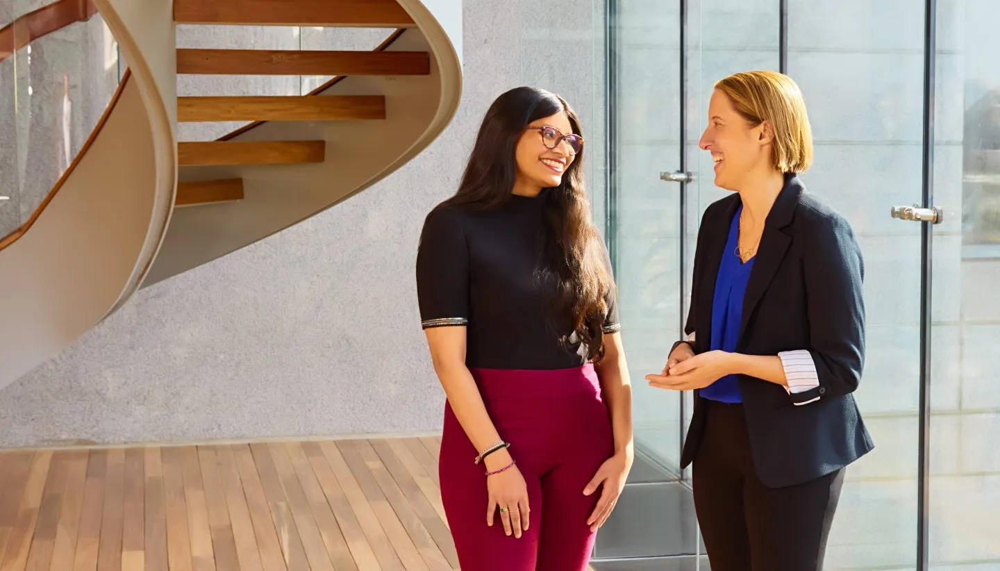 Two women smiling and talking in a sunlit modern space with a spiral staircase in the background