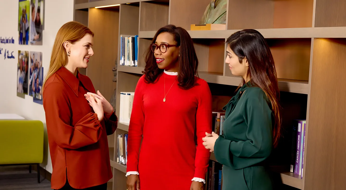 Three women engaged in a friendly conversation in front of bookshelves and a photo display wall