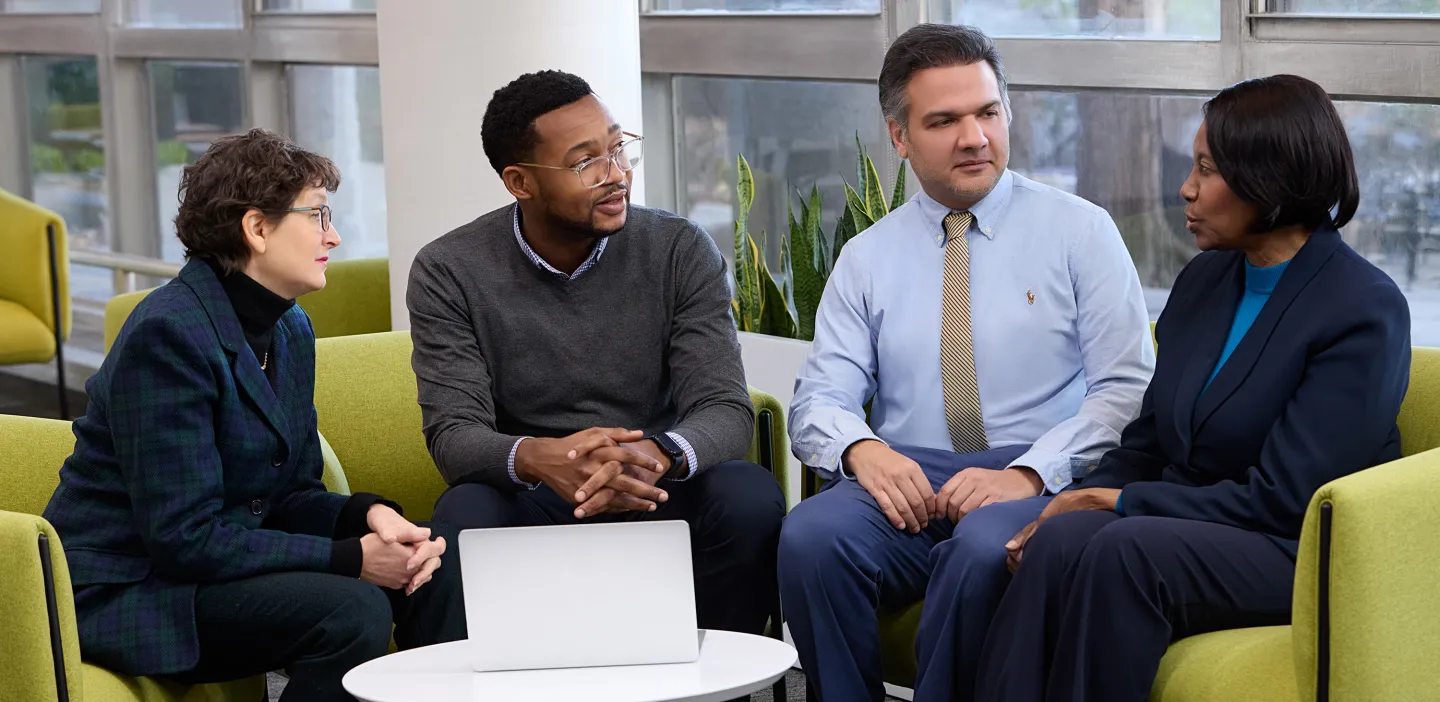 Four professionals sitting on green chairs having a serious discussion in a bright room with large windows