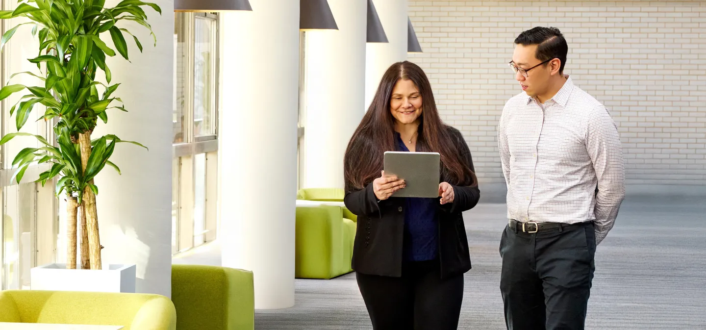 Two colleagues walking in a bright hallway, smiling while looking at a tablet together