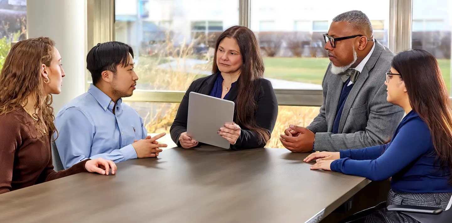 Five professionals seated around a table having a discussion, one person holding a tablet