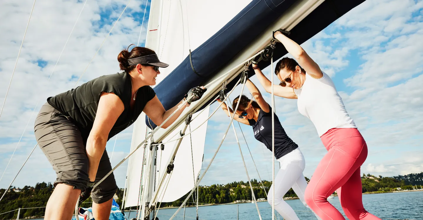 Three women working together to adjust the rigging on a sailboat under a sunny sky.