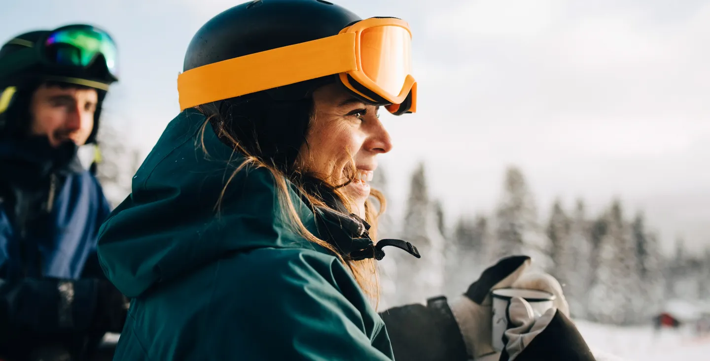 Woman in ski gear smiling with orange goggles on her helmet, standing outside in a snowy landscape with a friend in the background.