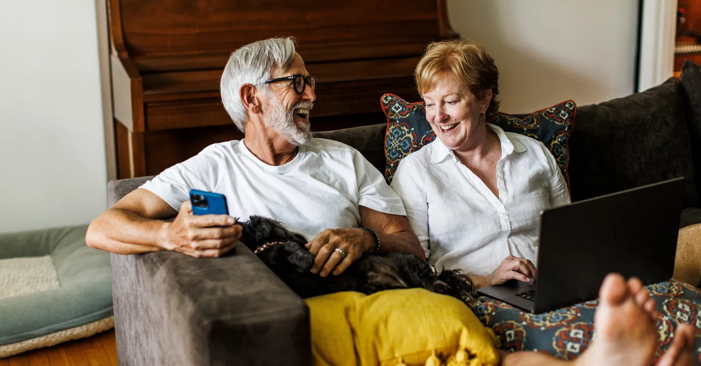 Older couple relaxing on a couch with a dog, smiling while using a smartphone and laptop.