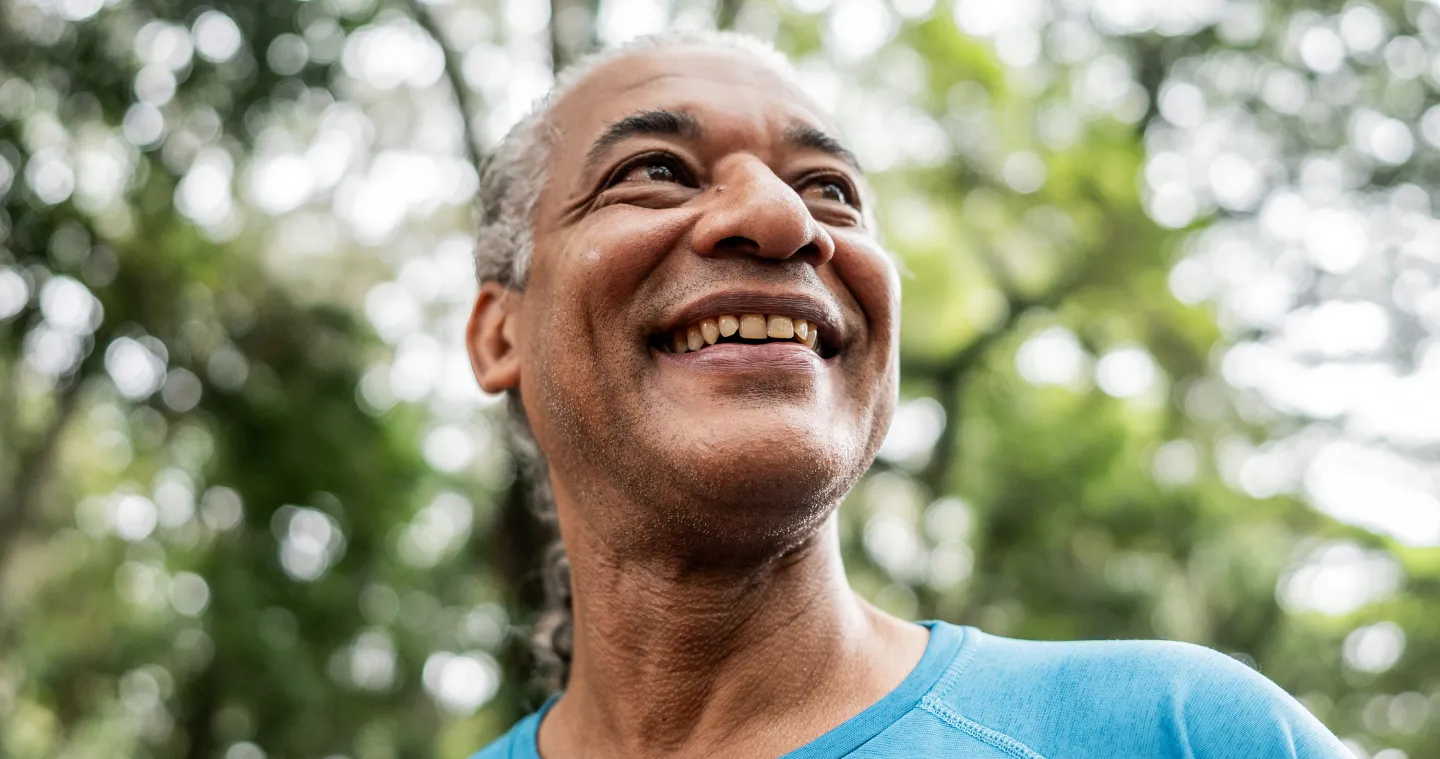 Close-up of a smiling man in a blue shirt outdoors, looking up with sunlight filtering through the trees behind him.