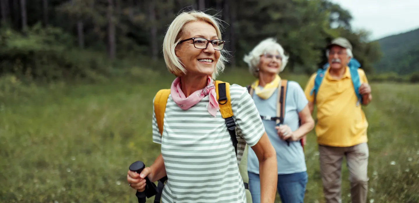 Group of older adults hiking through a grassy field with backpacks, led by a smiling woman in a striped shirt and pink scarf.