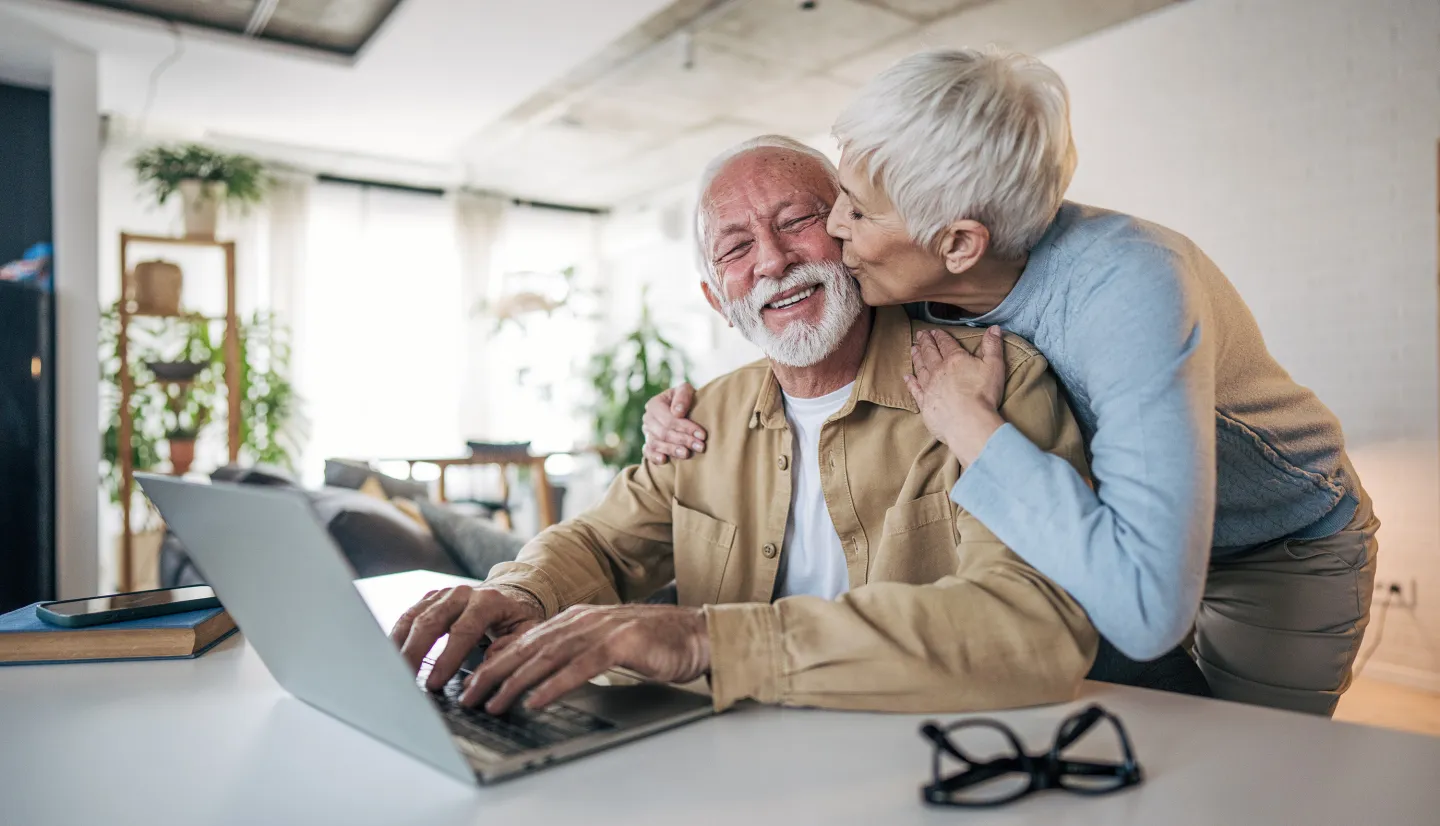Smiling senior man using a laptop while his partner kisses him on the cheek in a bright, cozy home setting.