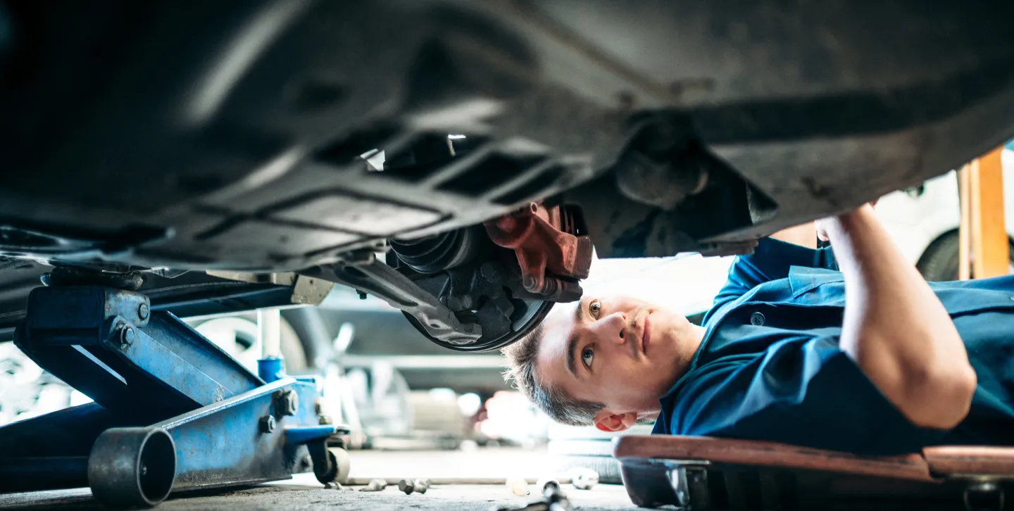 Male mechanic lying on a creeper and inspecting the underside of a vehicle in an auto repair shop.