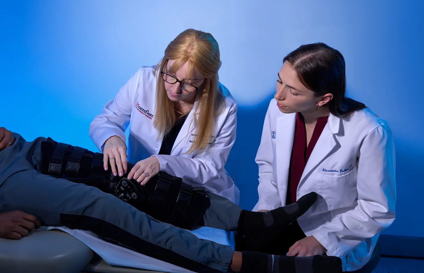 Physician adjusting a knee brace on a patient’s leg while a medical student observes attentively in a clinical setting.