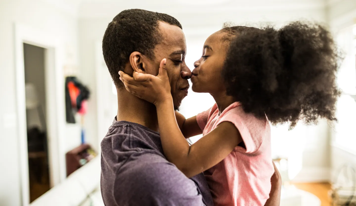 Young girl kissing her father's nose while being held in his arms, both smiling in a warmly lit home setting.