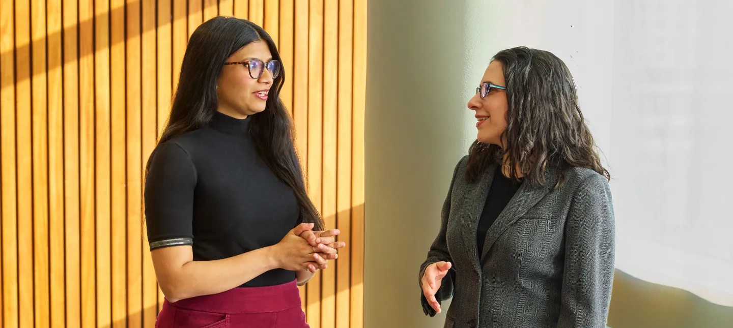 Two women standing and smiling while having a conversation near a window and wood-paneled wall.