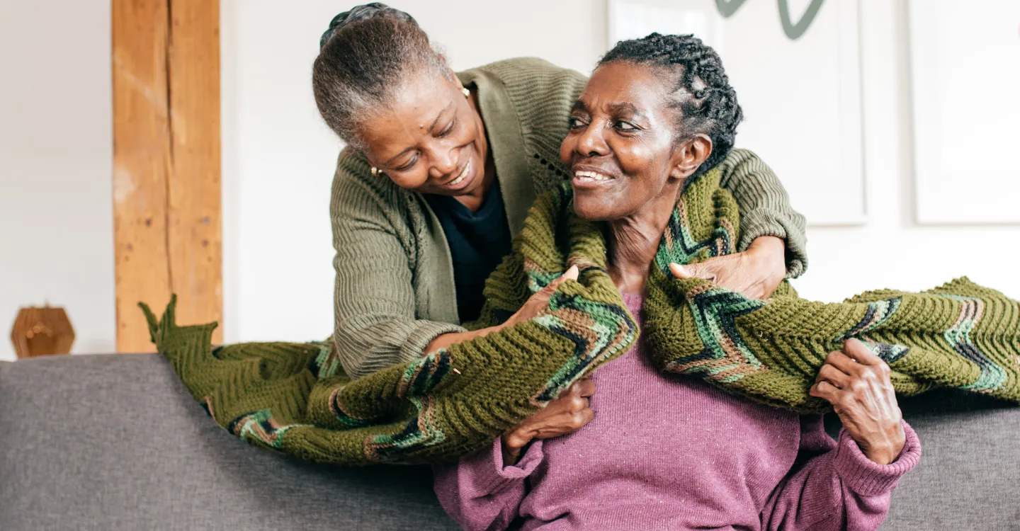 Smiling woman drapes warm knitted blanket over older woman on couch