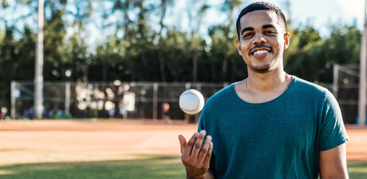 Young man smiling and tossing a baseball on a field, standing in front of a batting cage.