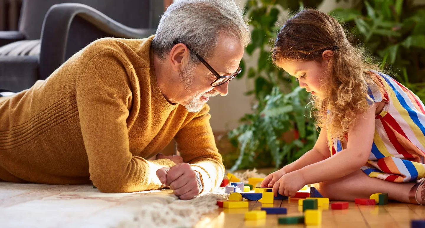 Smiling man lying on the floor playing with colorful wooden blocks alongside a young girl in a striped dress.