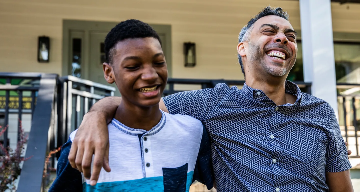 Smiling man with his arm around a laughing teenage boy as they stand outside a house on a sunny day.