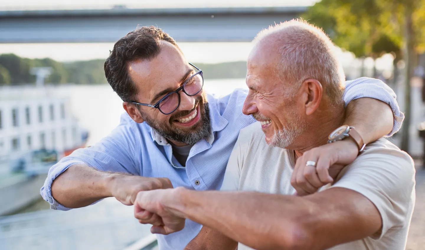Two men smiling and greeting each other with an elbow bump near a waterfront.