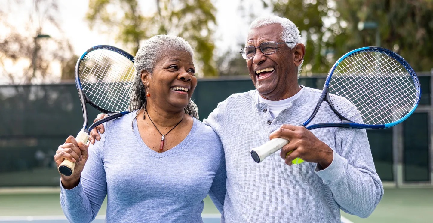 Smiling older couple holding tennis rackets and embracing on a court, enjoying a sunny day together.