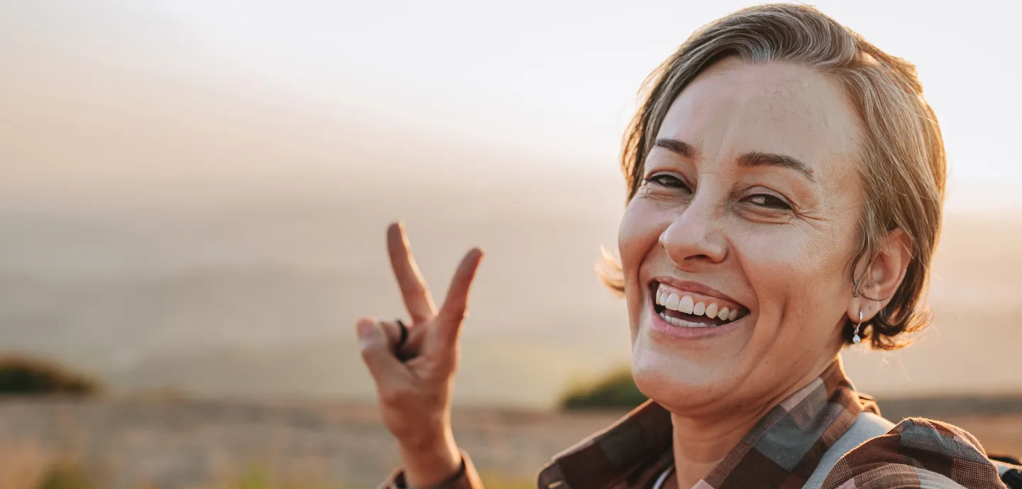 Smiling woman flashing a peace sign while standing outdoors at sunset.