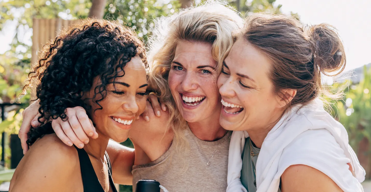 Three women smiling and embracing outdoors on a sunny day, sharing a joyful moment together.