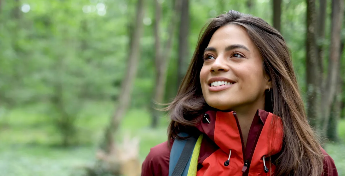 Smiling woman in a red jacket and backpack looking up while walking through a lush green forest.