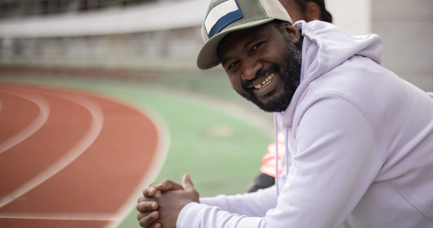 Man wearing a white hoodie smiles while sitting beside a track field.