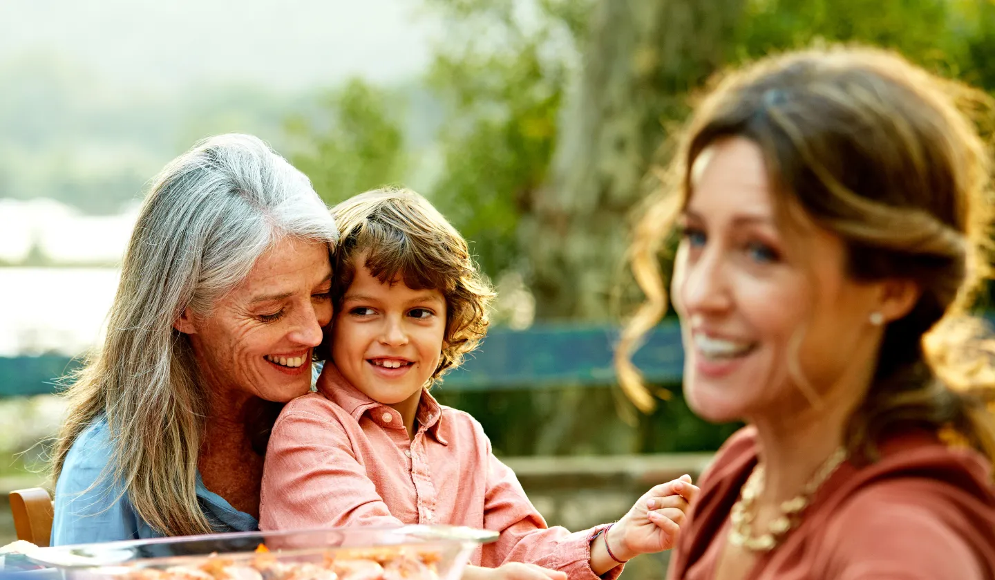 Smiling older woman hugging a young boy outdoors with another woman in the foreground, all enjoying a sunny day together.