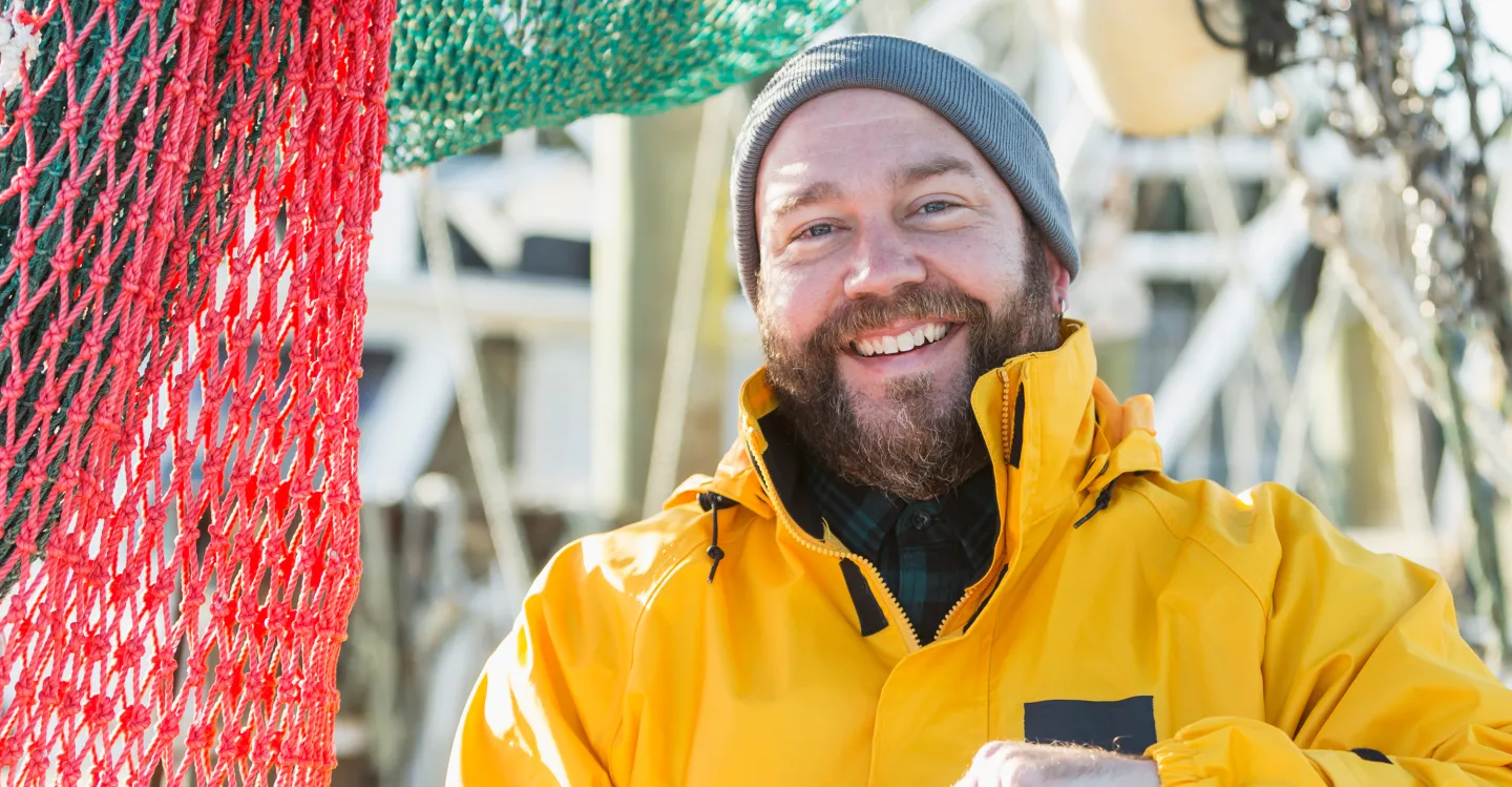 Smiling man in a yellow jacket and gray beanie standing next to colorful fishing nets in a sunny harbor setting.