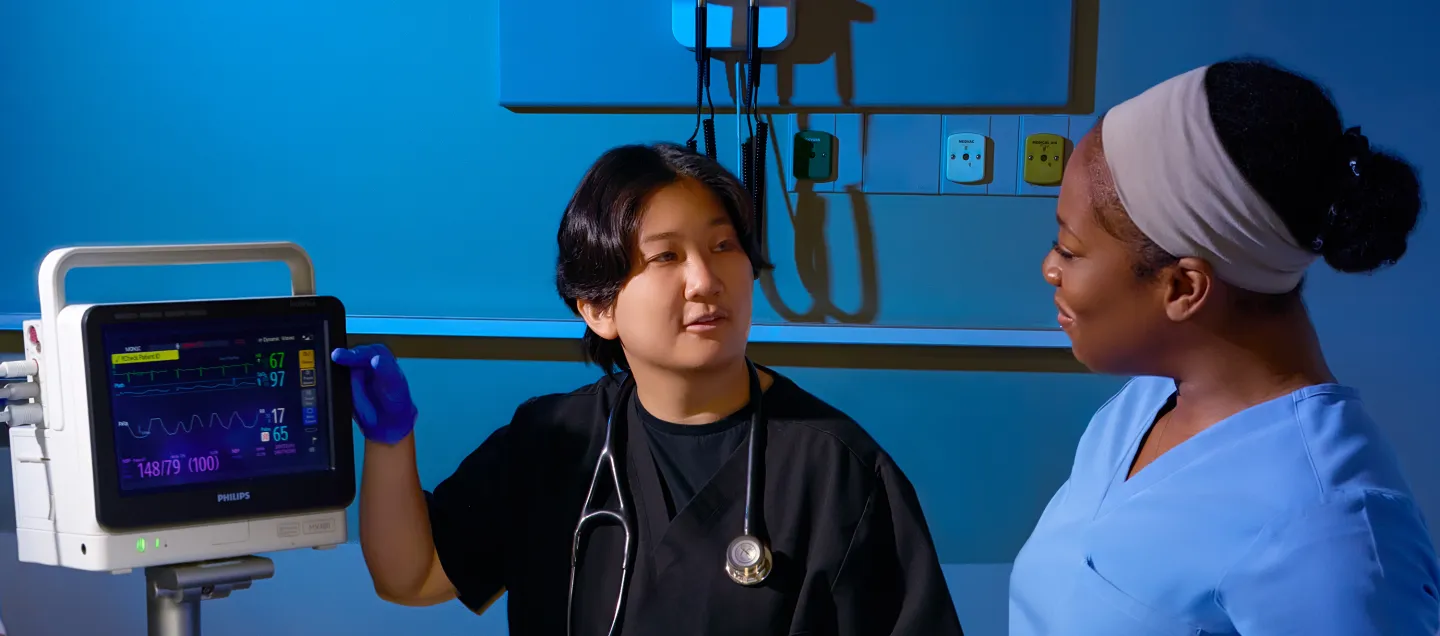 Nurse practitioner explains patient monitor readings to a fellow healthcare professional in a clinical setting, demonstrating collaborative care and medical training.