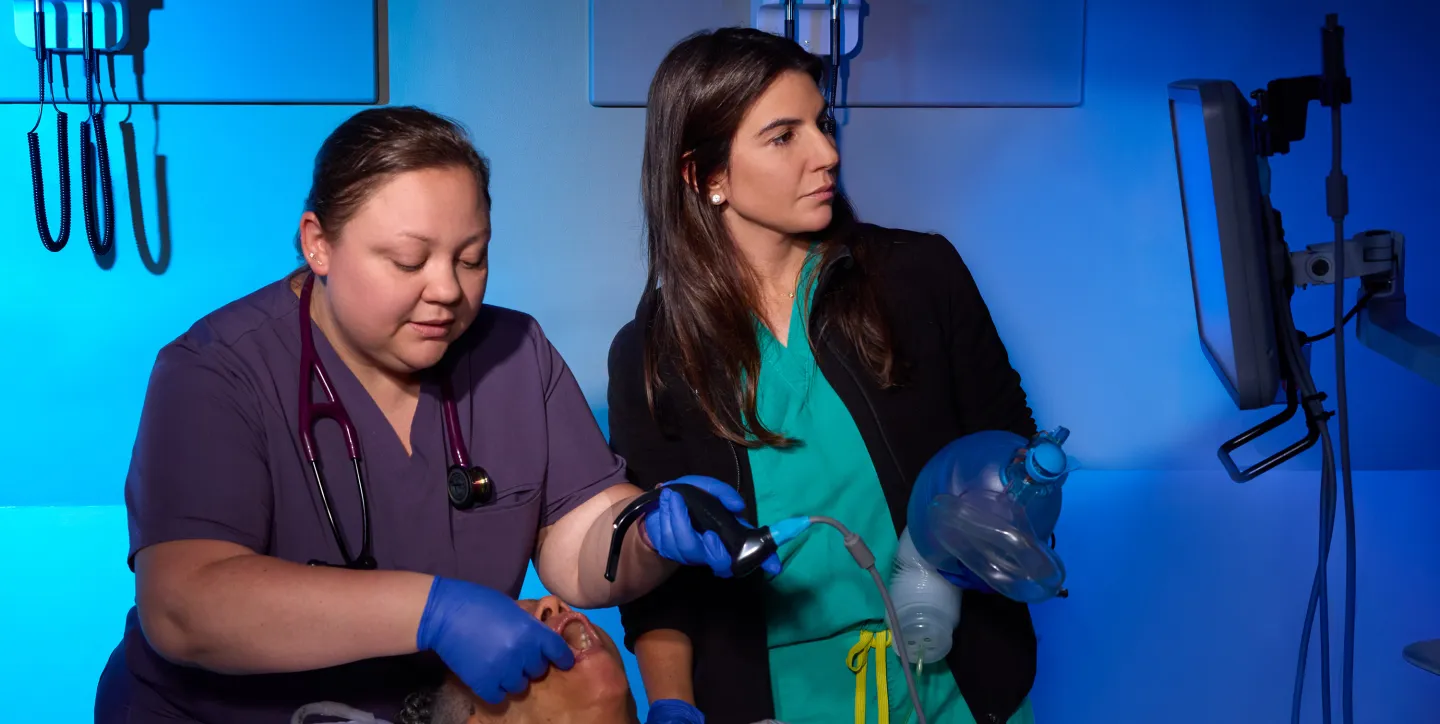 A nurse and a medical instructor perform a simulated airway procedure on a mannequin, using a laryngoscope and bag valve mask, under blue-toned clinical lighting.