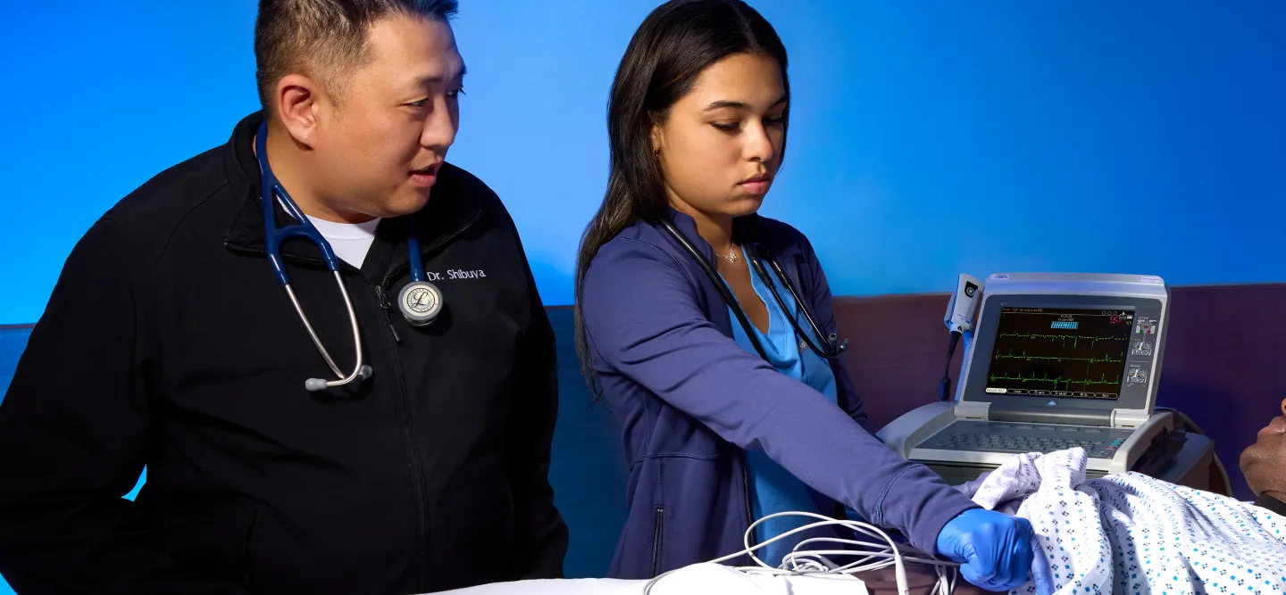 A medical fellow monitors a patient while another healthcare provider performs an EKG test on a hospital bed, showcasing emergency medicine residency training.