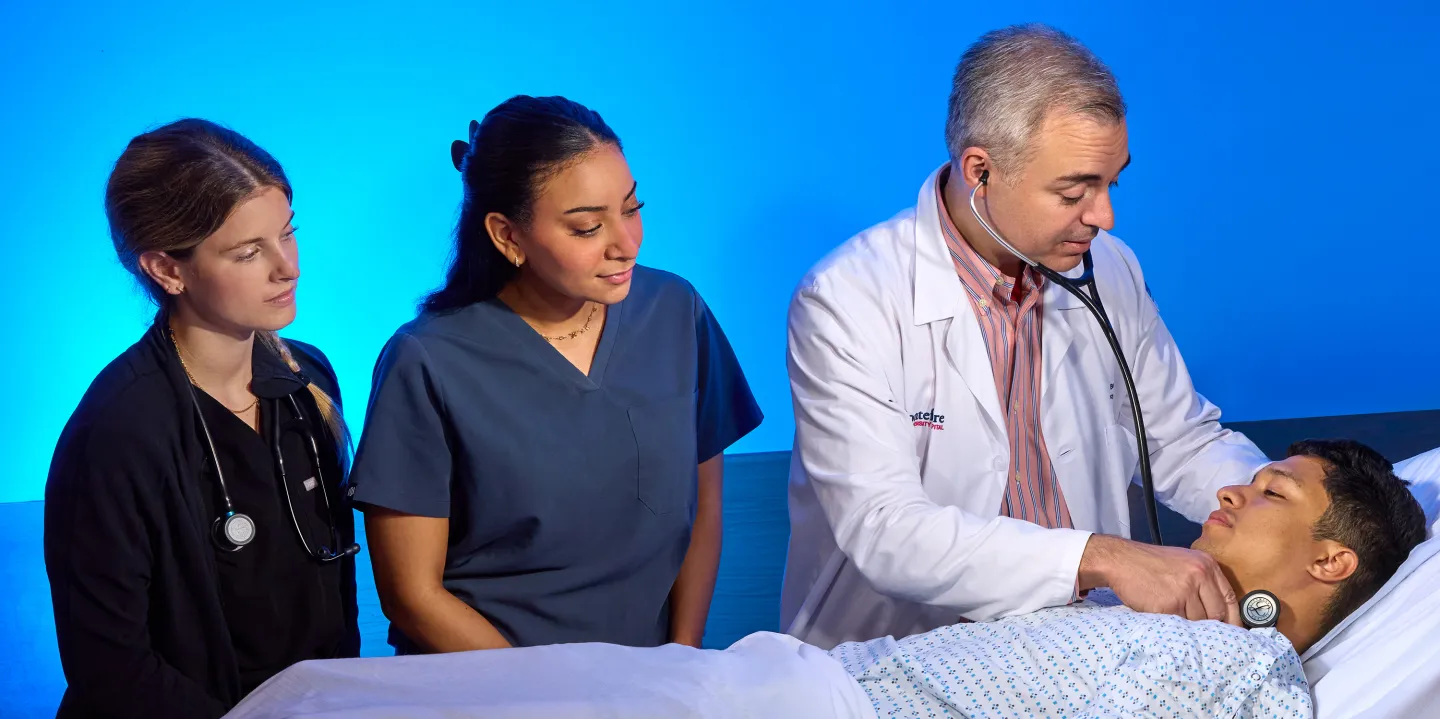 A doctor in a white coat uses a stethoscope on a patient in a hospital bed while two medical students observe closely.