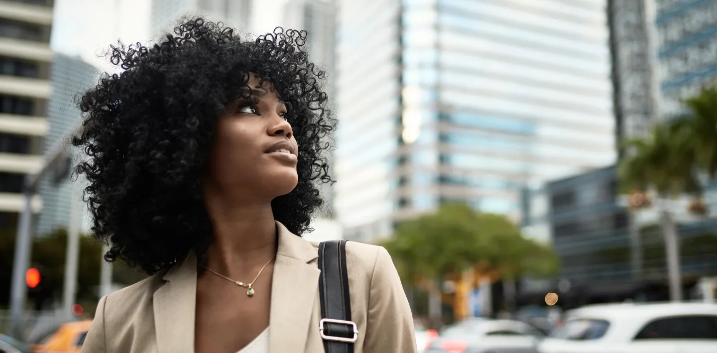 Confident woman with curly hair looking upward while walking through a city street, with modern glass skyscrapers in the background.