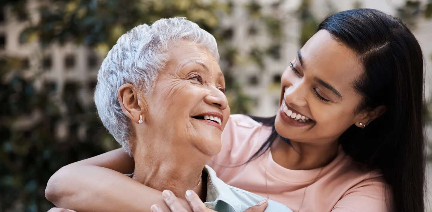 Smiling older woman being embraced by a younger woman, both sharing a joyful moment outdoors.