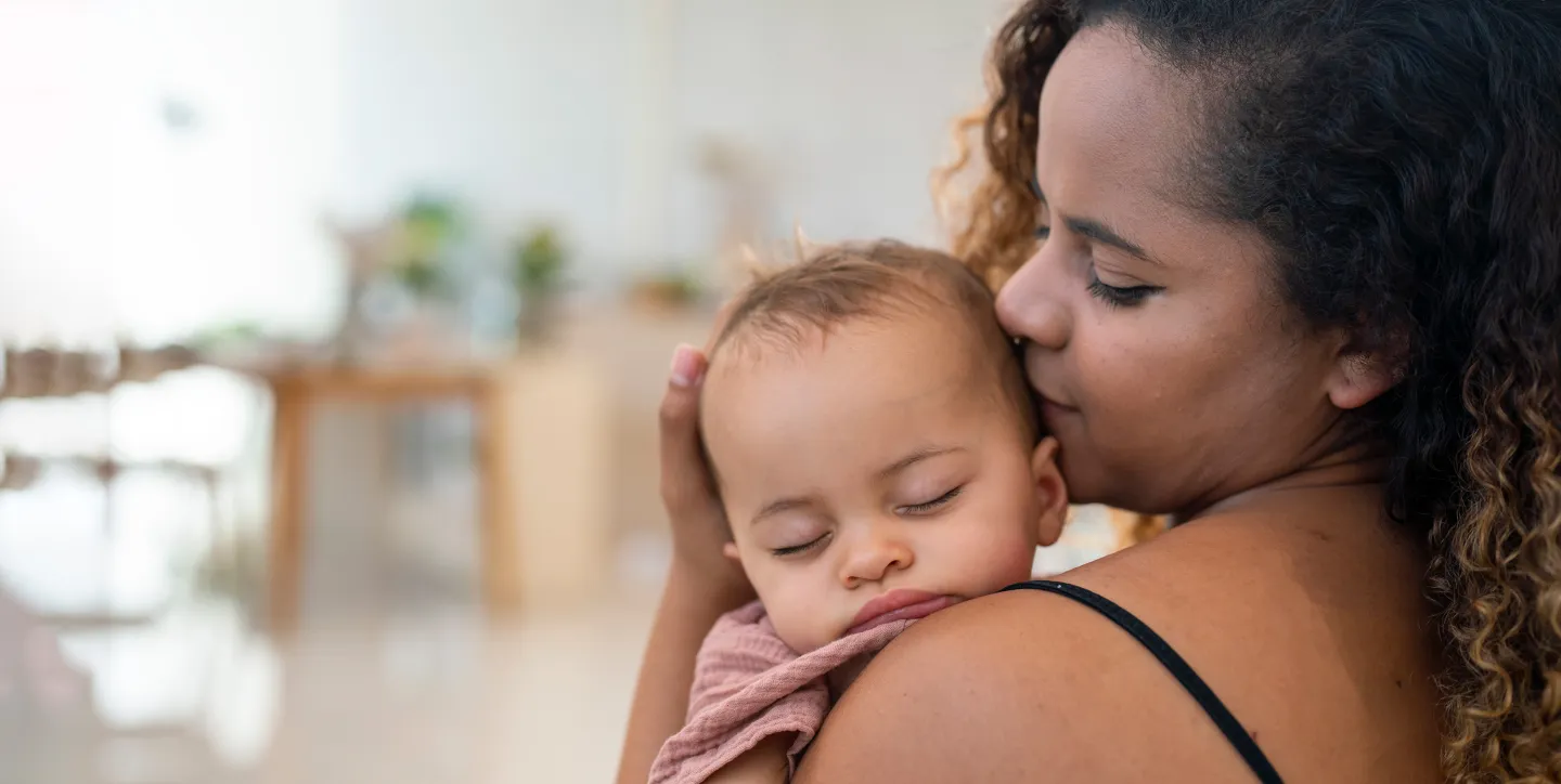 Mother gently holding and kissing her sleeping baby against her shoulder in a softly lit home environment.