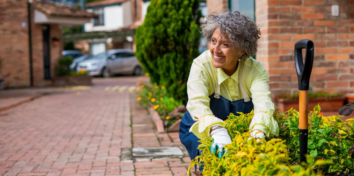 Smiling older woman wearing gardening gloves and a yellow shirt, tending to a vibrant flower bed in a brick courtyard.