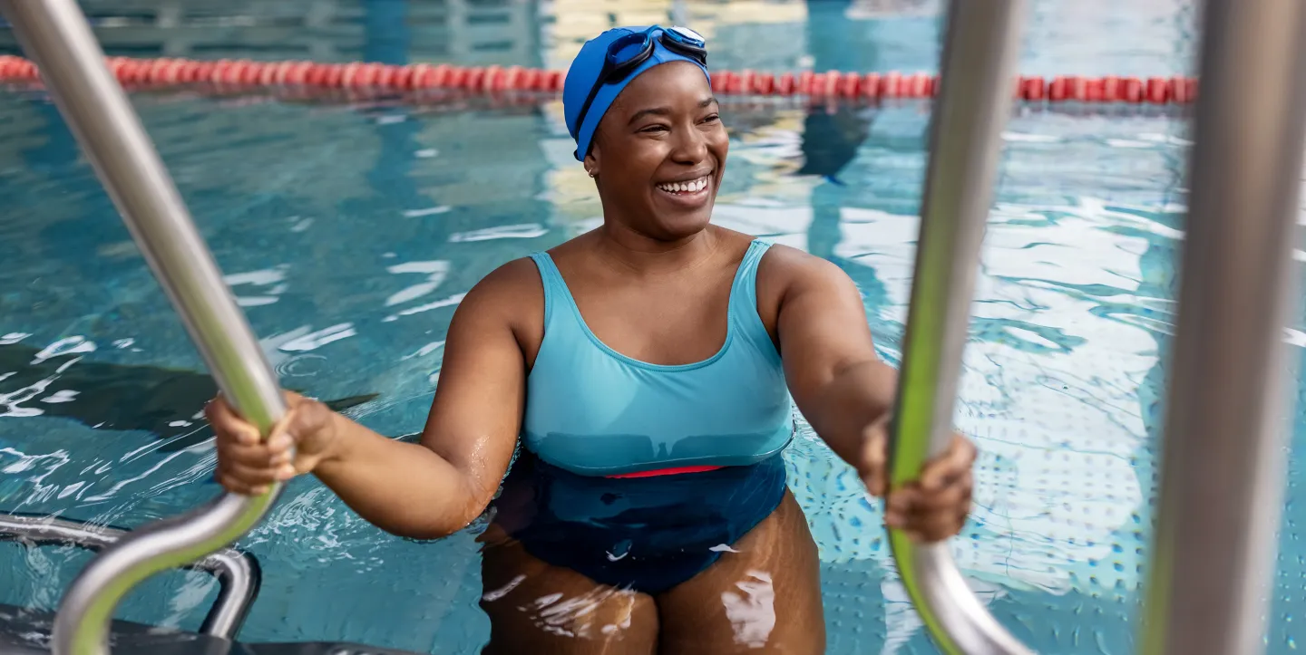 Smiling woman in a blue swimsuit and swim cap holding onto pool handrails as she exits the water at an indoor swimming facility.