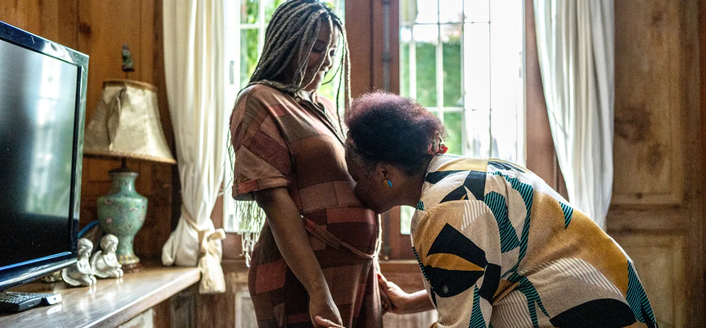 A woman kneels and kisses the belly of a pregnant woman standing in a warmly lit home with vintage furniture and large curtained windows.