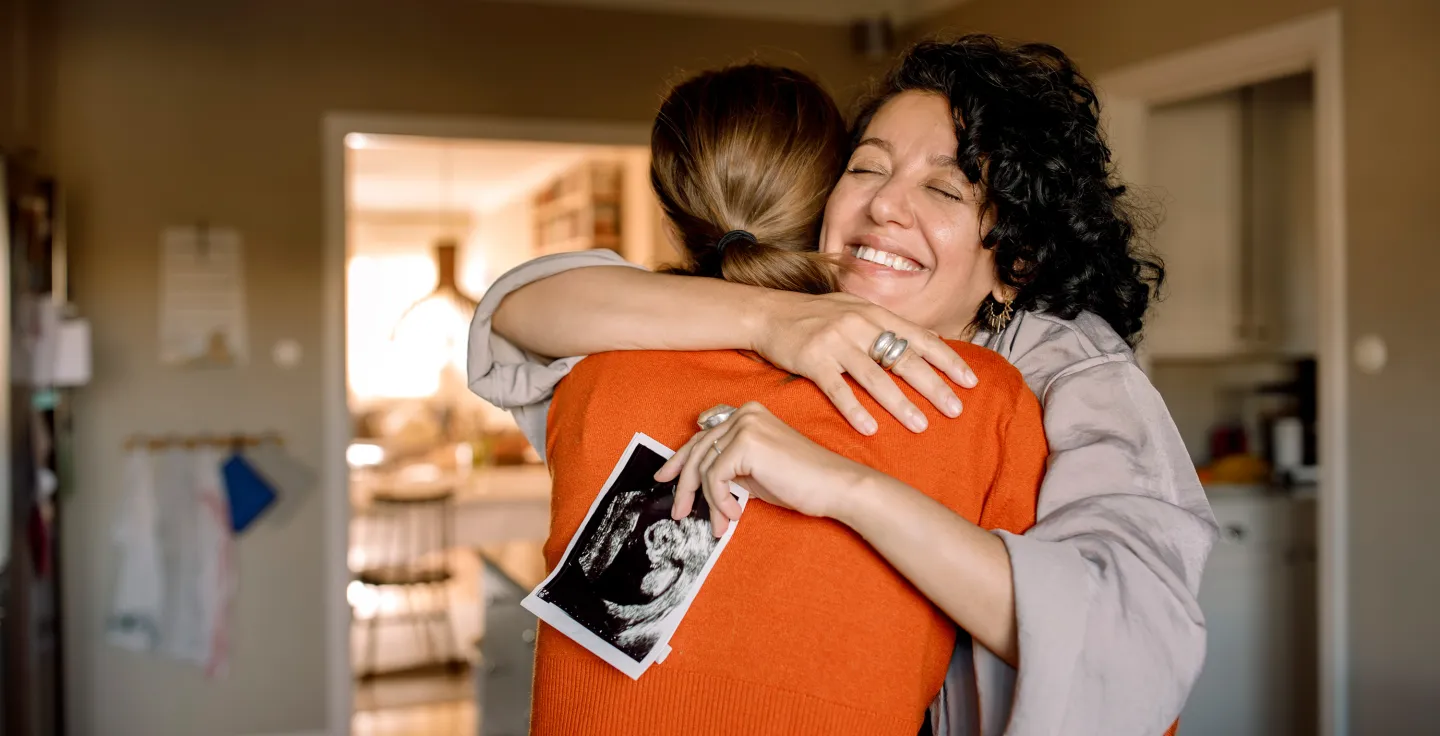 Joyful moment between two women hugging in a warmly lit kitchen, one of them holding a printed ultrasound image against her back.